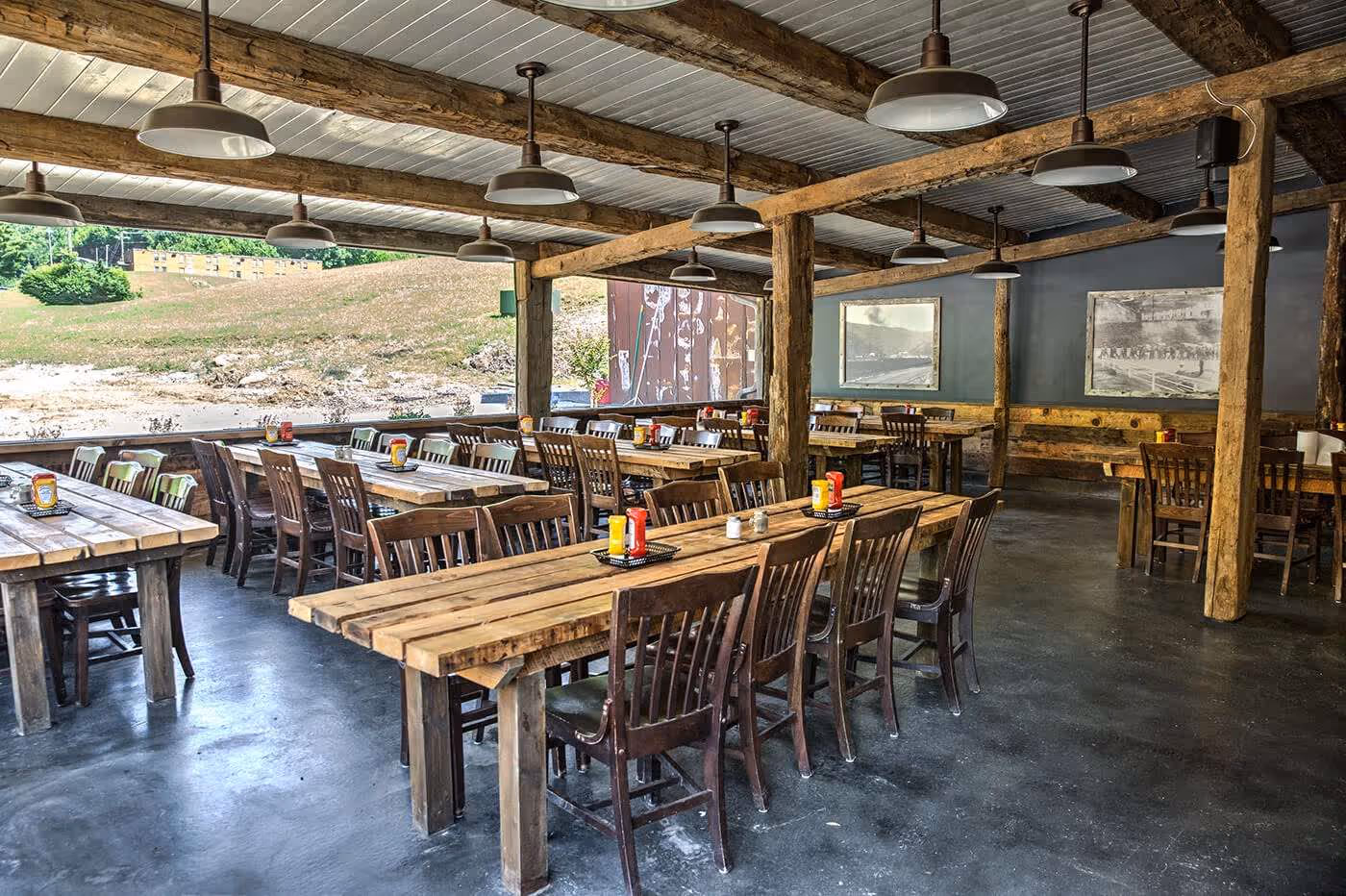 Rustic restaurant interior with wooden picnic-style tables and chairs, condiment bottles on tables, and a view of grassy outdoor area through open side.