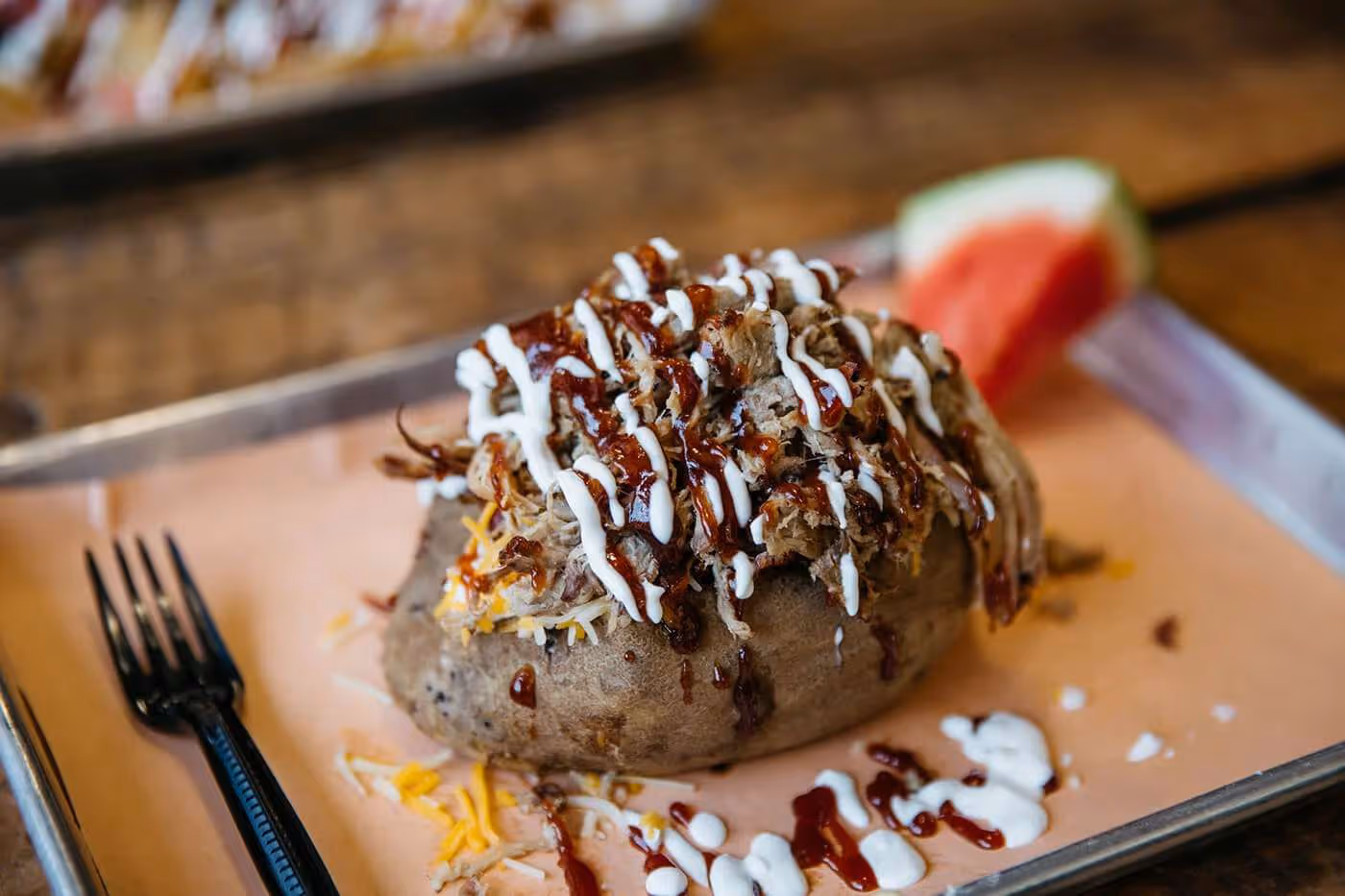 Baked potato topped with shredded cheese, pulled pork, and drizzled with barbecue and sauces on a metal tray with a watermelon slice and a black fork.