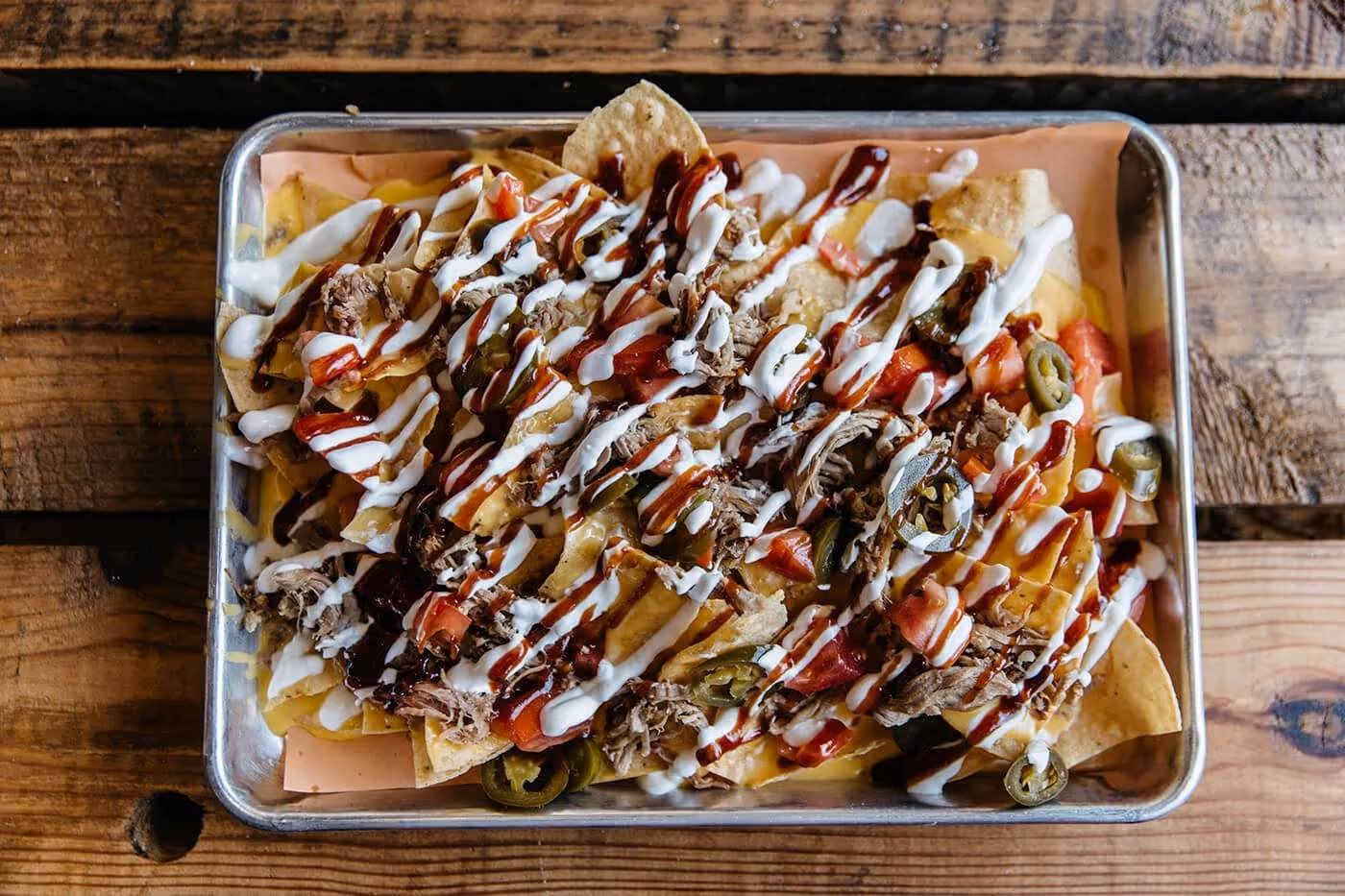 Tray of loaded nachos topped with shredded meat, diced tomatoes, jalapeños, cheddar cheese, sour cream, and barbecue sauce on a wooden table.