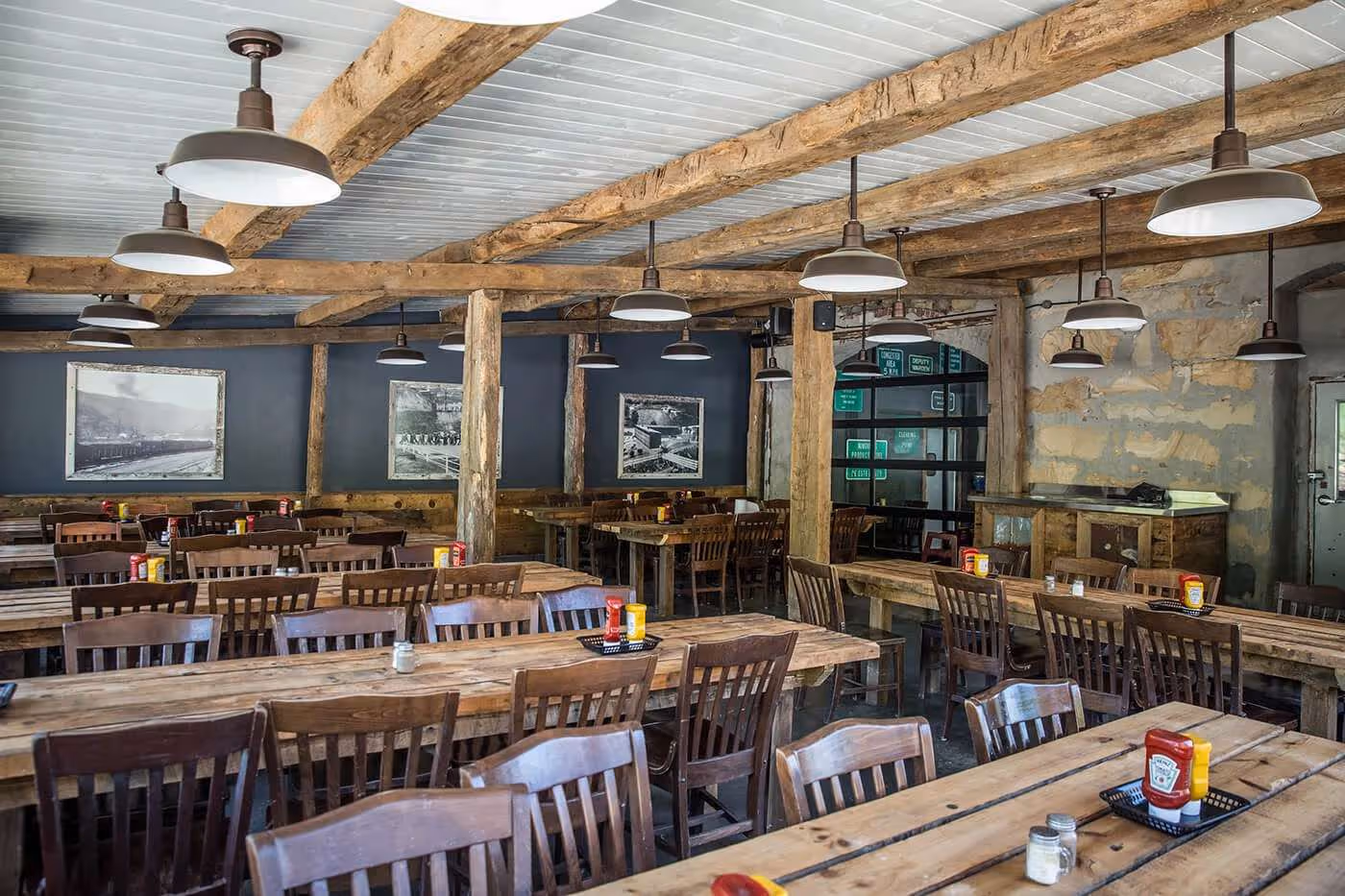 Rustic restaurant interior with wooden tables and chairs, exposed wooden beams, hanging industrial light fixtures, and condiment holders on tables.
