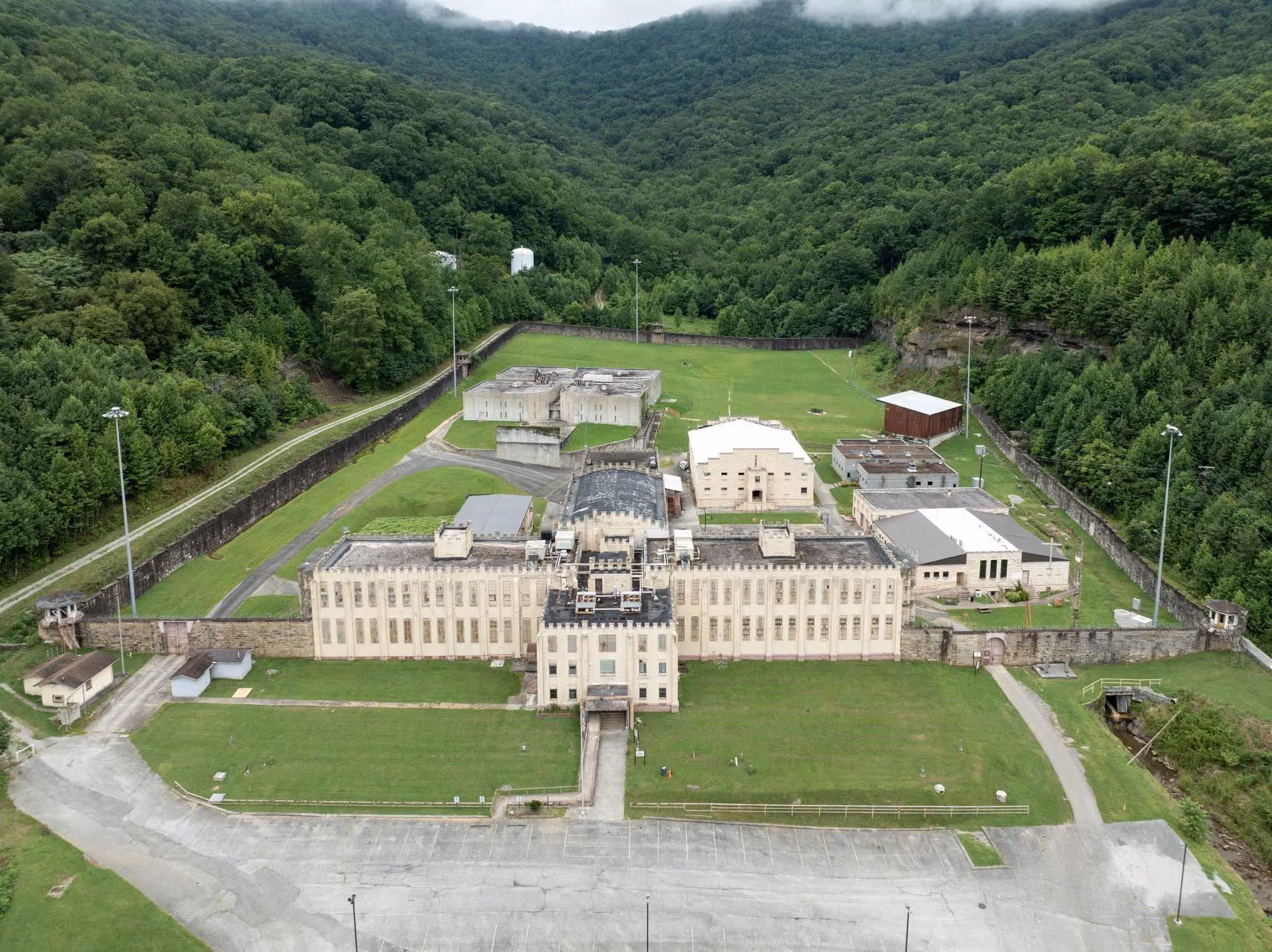 Aerial view of a large, historic prison complex surrounded by green forested hills and tall fences.