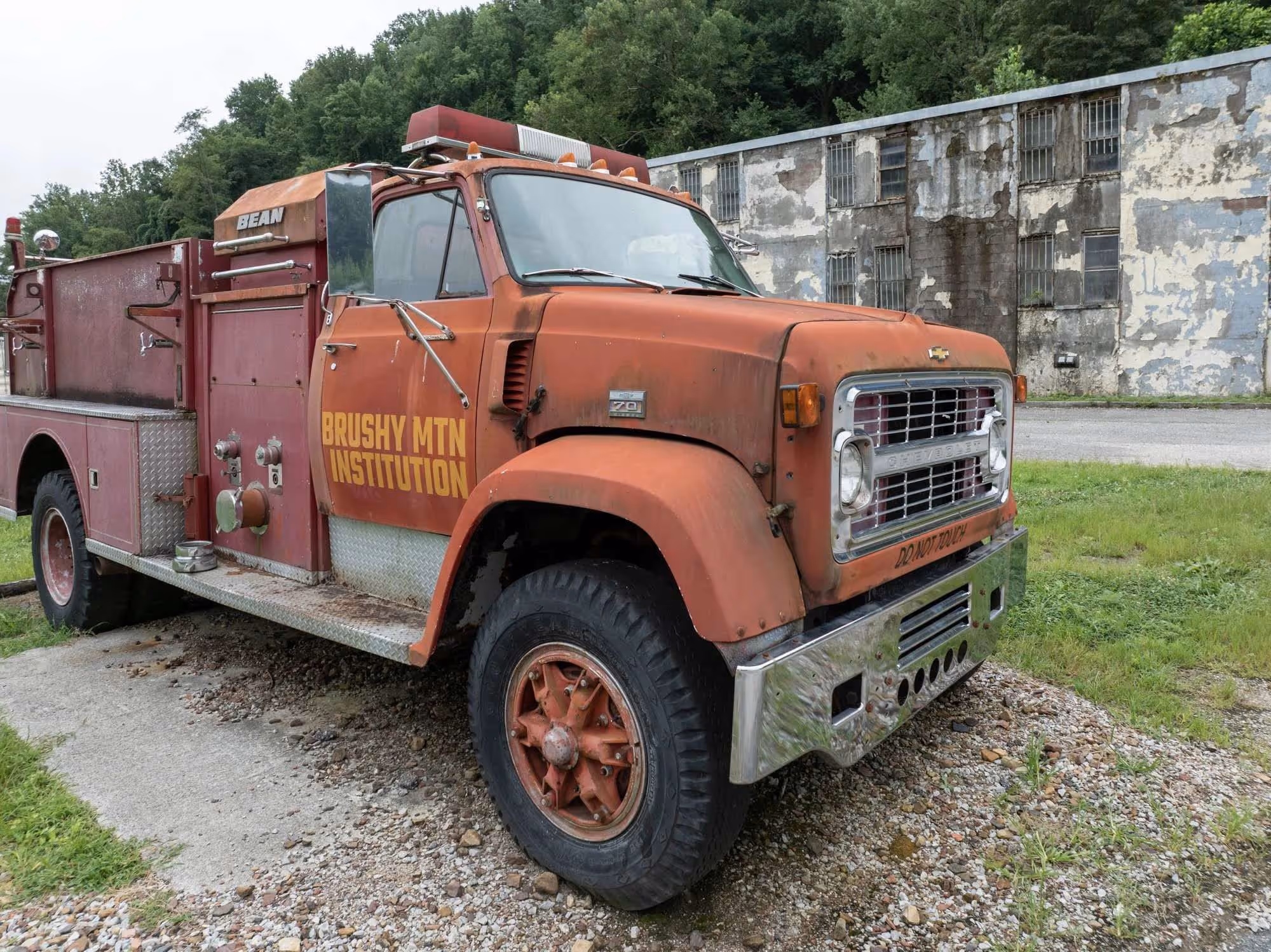 Rusty red Brushy Mountain Institution fire truck parked on gravel near an old building.