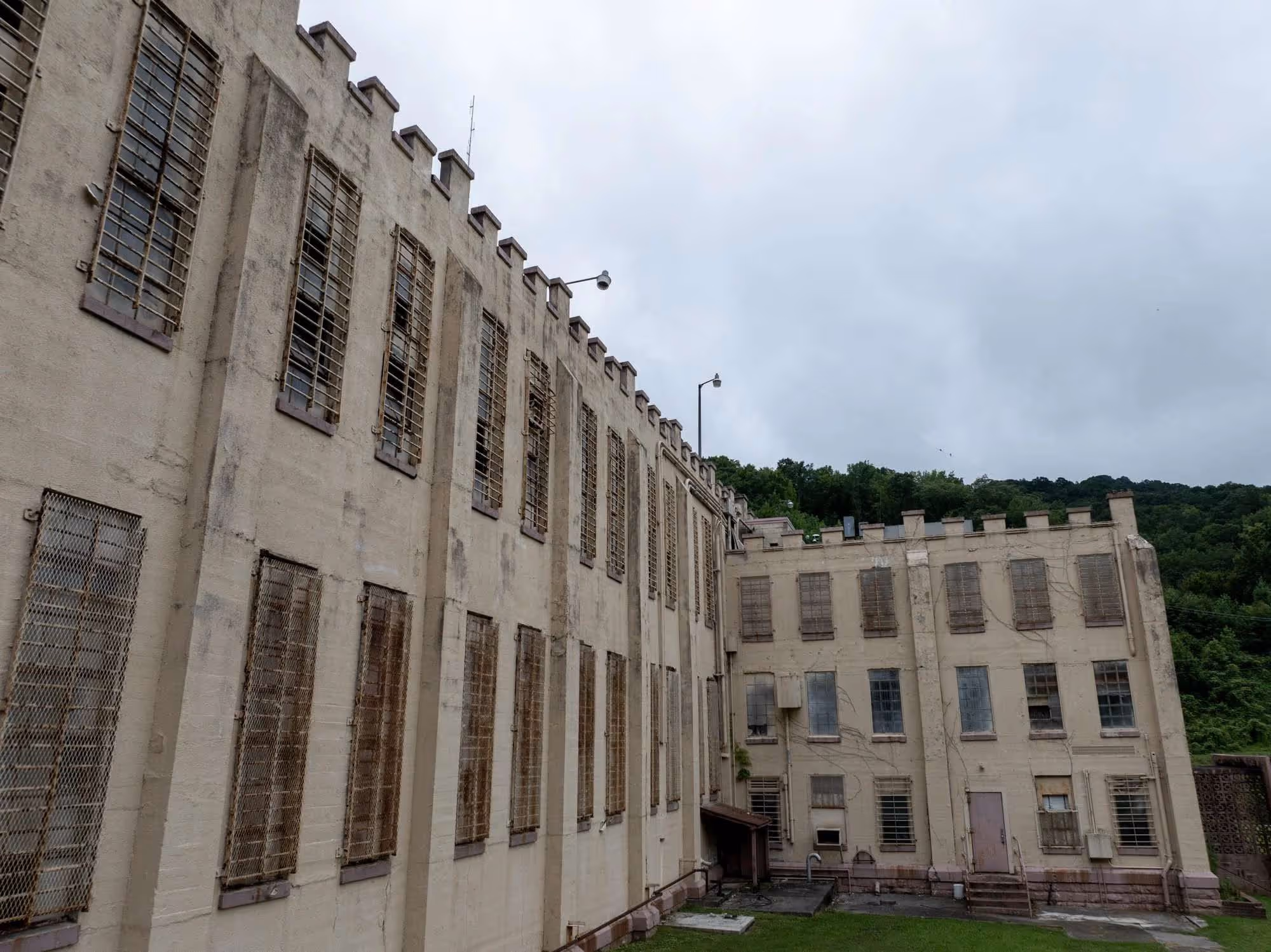 Exterior of a large, old beige prison building with barred windows and a green wooded hillside in the background under a cloudy sky.