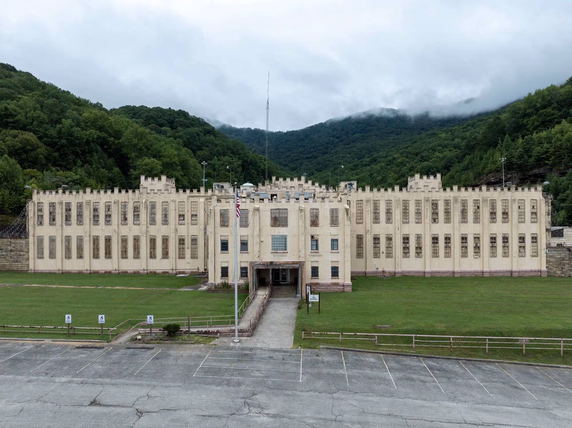 Large, beige, castle-like prison building with tall, barred windows, situated in front of green forested hills under a cloudy sky.