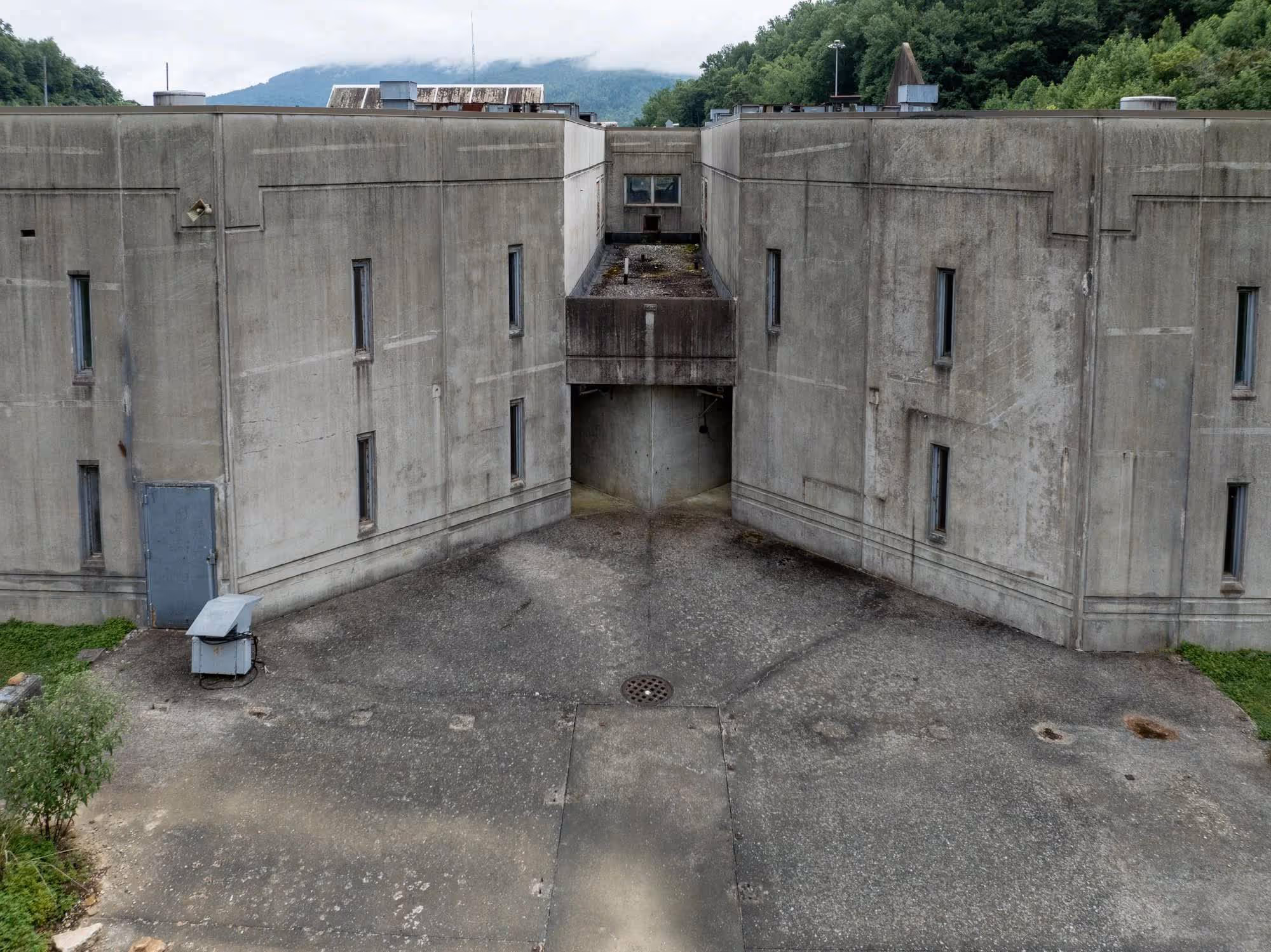 Concrete walls forming a narrow, enclosed courtyard with a small door and windows, surrounded by greenery and mountains in the background.