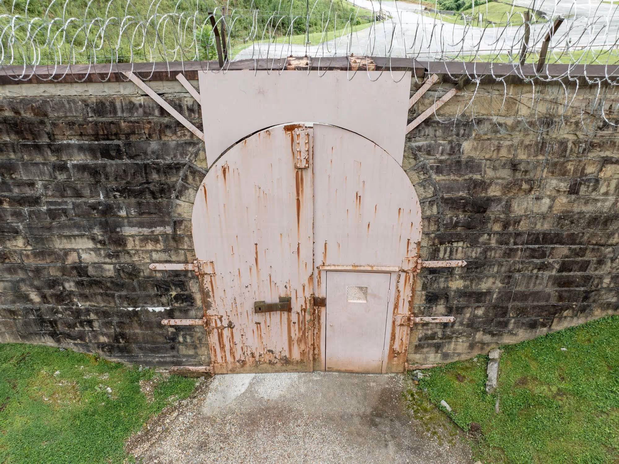 Rusty metal prison door with a smaller locked door inset, set in a stone wall topped with barbed wire.