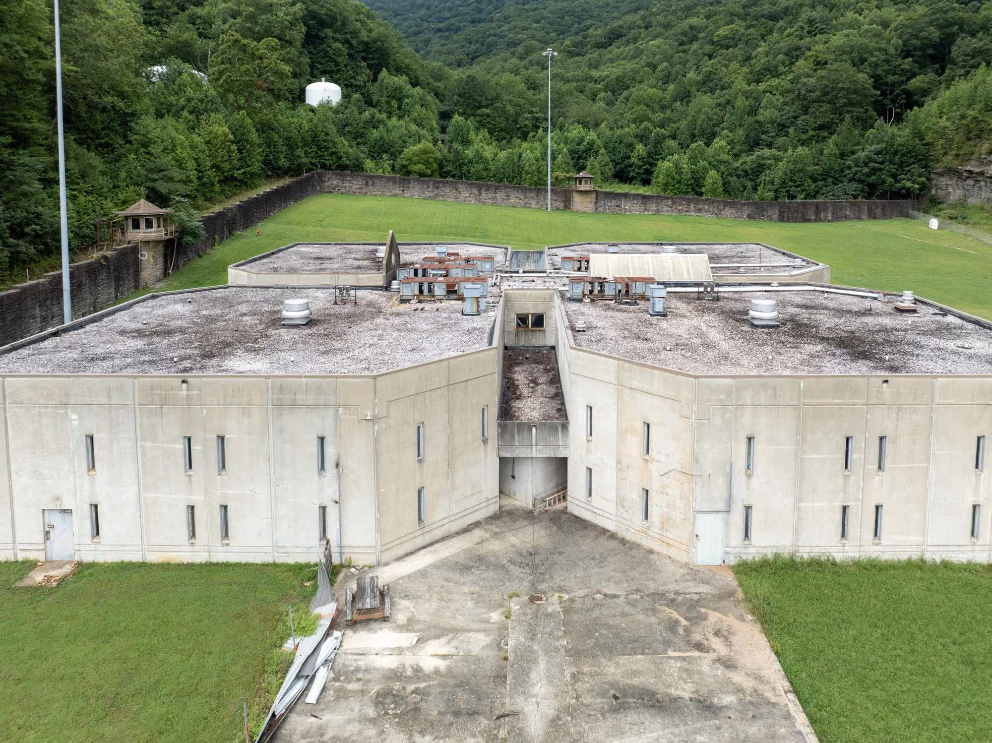 Aerial view of a large concrete prison building surrounded by tall stone walls and watchtowers, with green forested hills beyond.