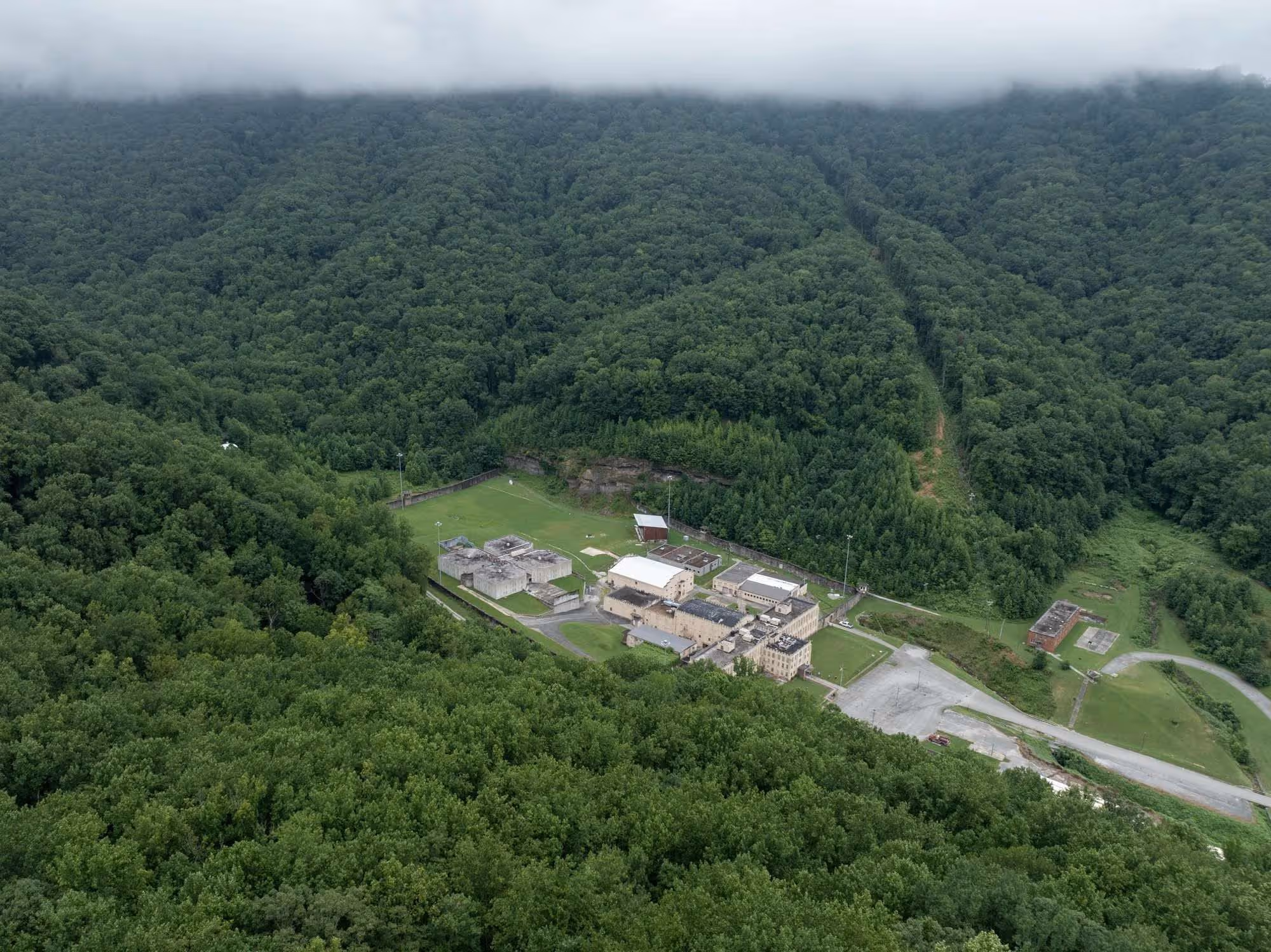 Aerial view of the prison complex surrounded by dense green forest and hills with low clouds.