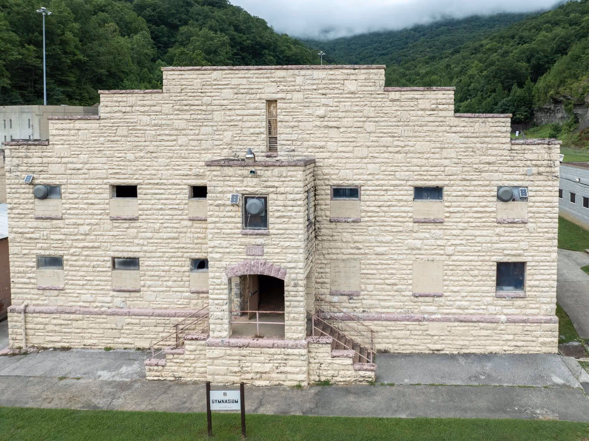 Old stone gymnasium building with boarded-up windows, stairs leading to an arched entrance, and lush green hills in the background.