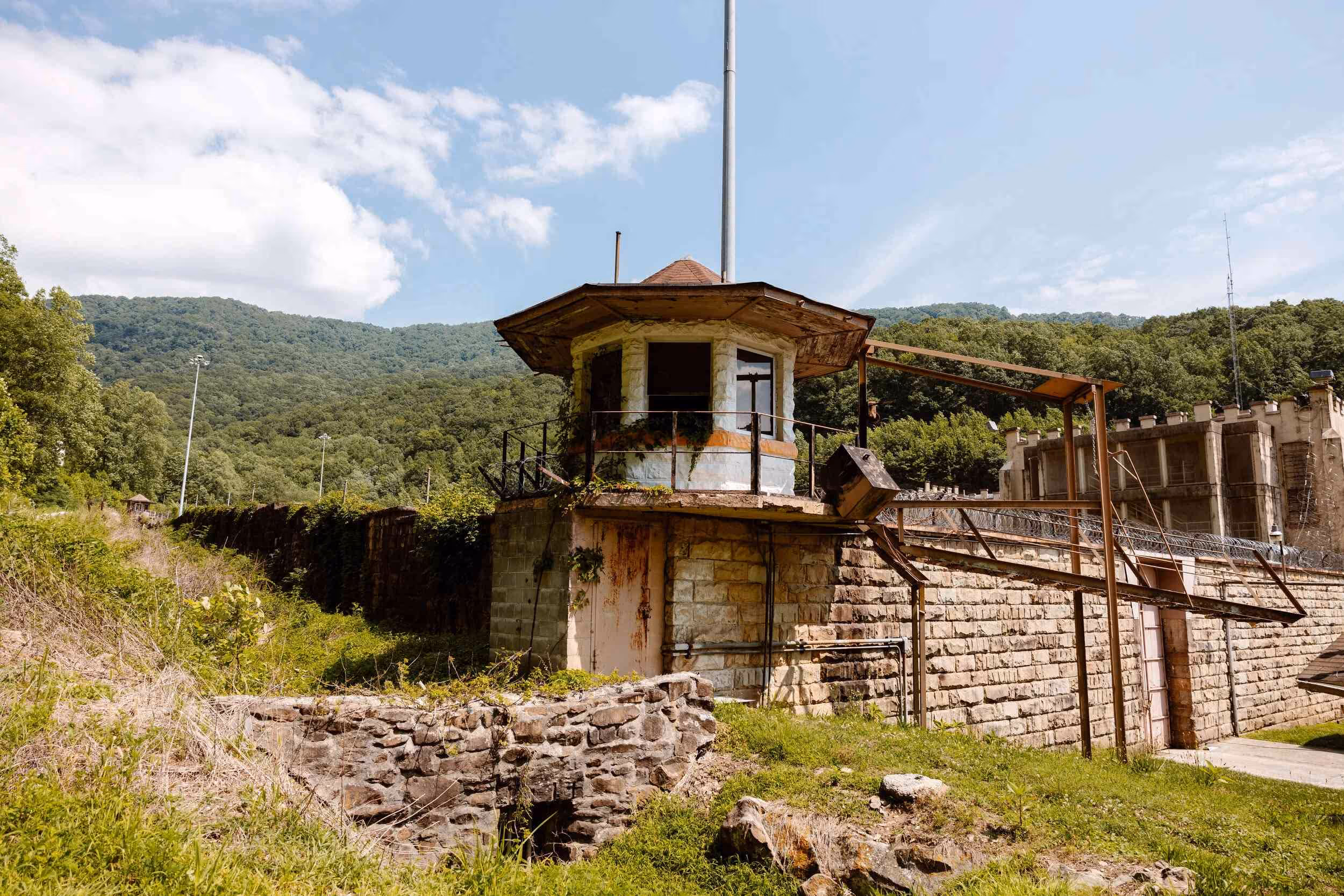 Old stone guard tower with metal railing and rusty door situated on a grassy hill with forested mountains in the background under a partly cloudy sky.