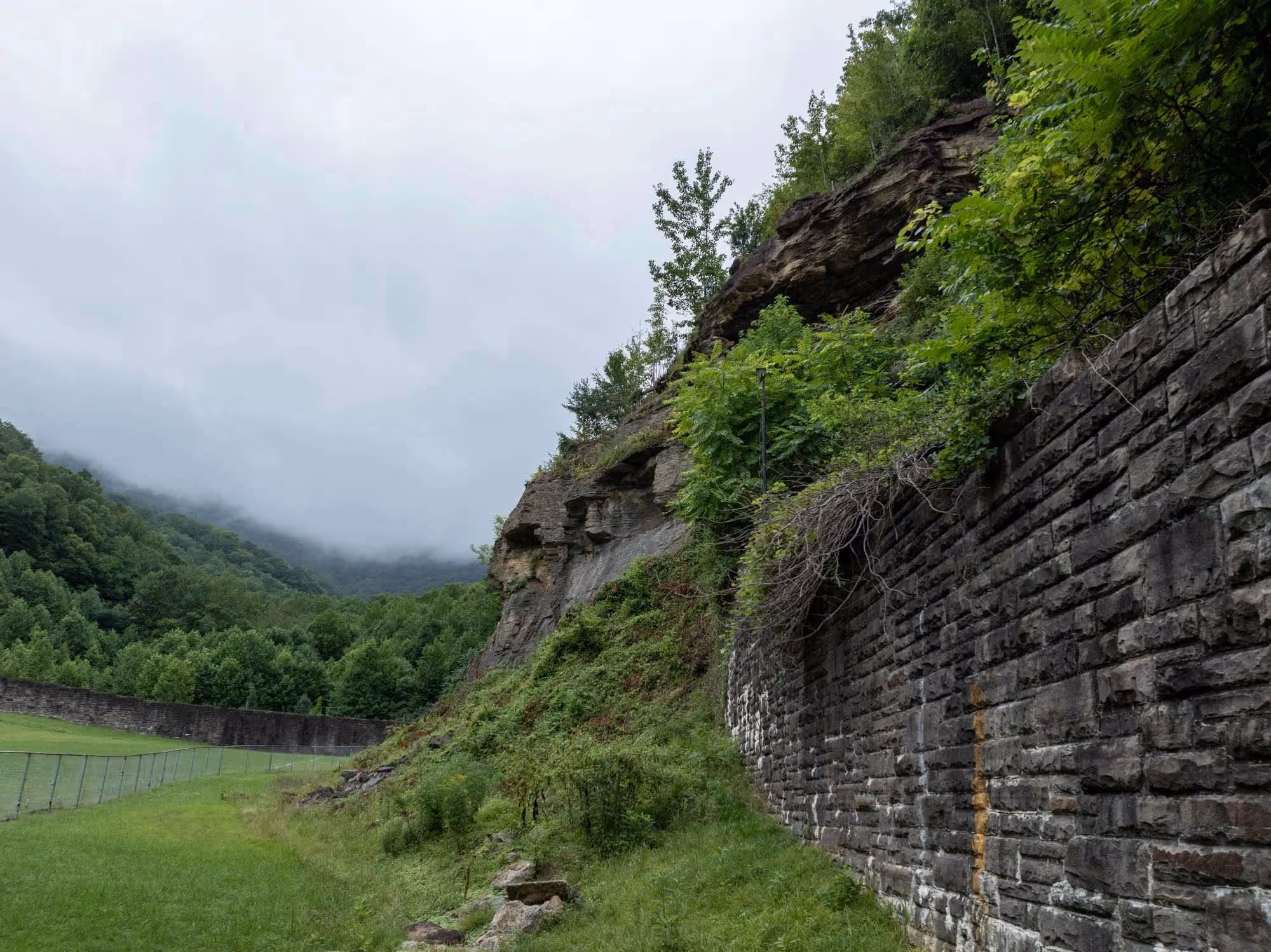 Foggy mountain landscape with green trees and rocky cliff beside a grassy field with a fence and the stone prison wall.