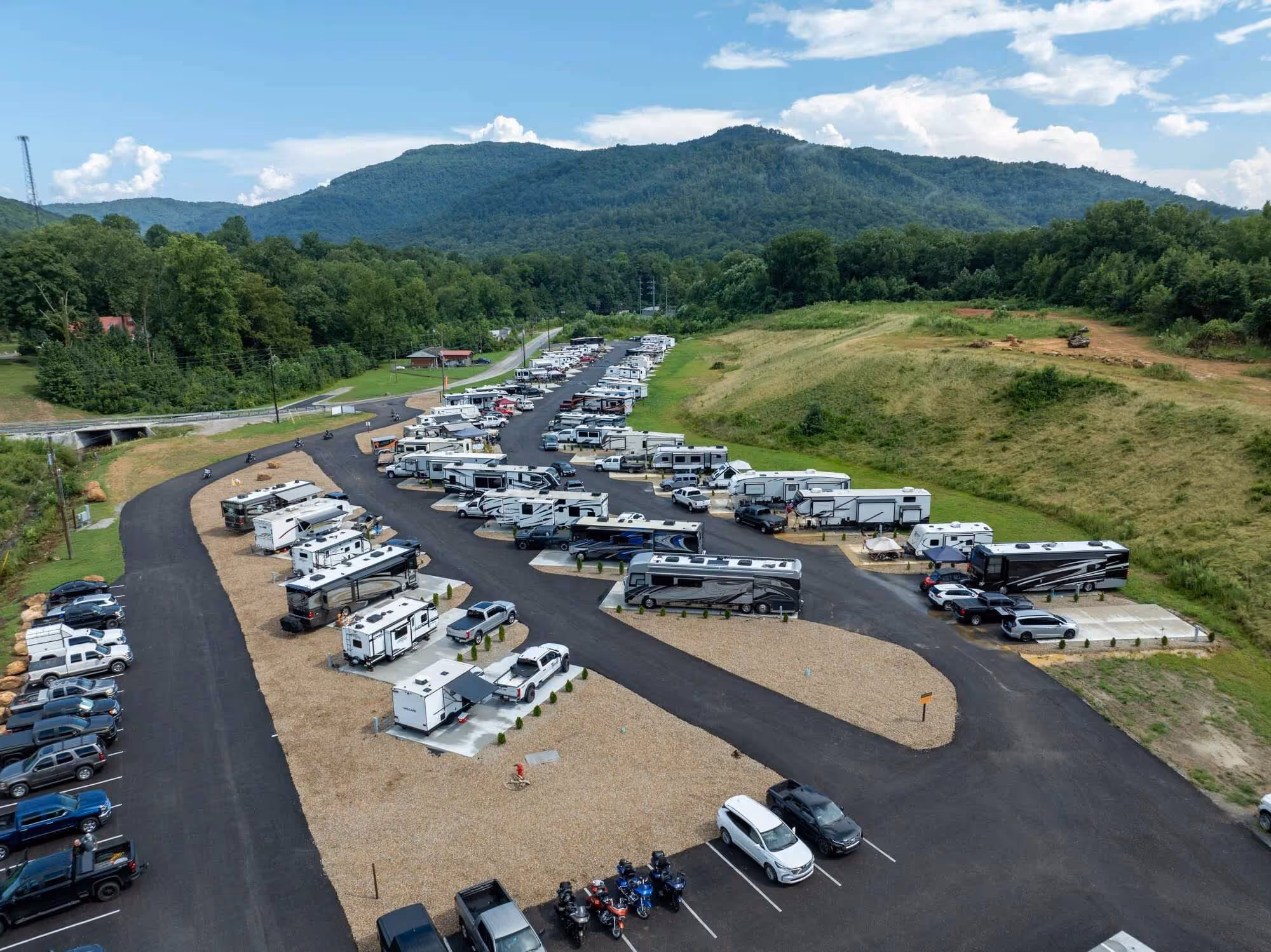 Aerial view of Brushy RV park with multiple parked recreational vehicles and cars, surrounded by green trees and hills under a partly cloudy sky.