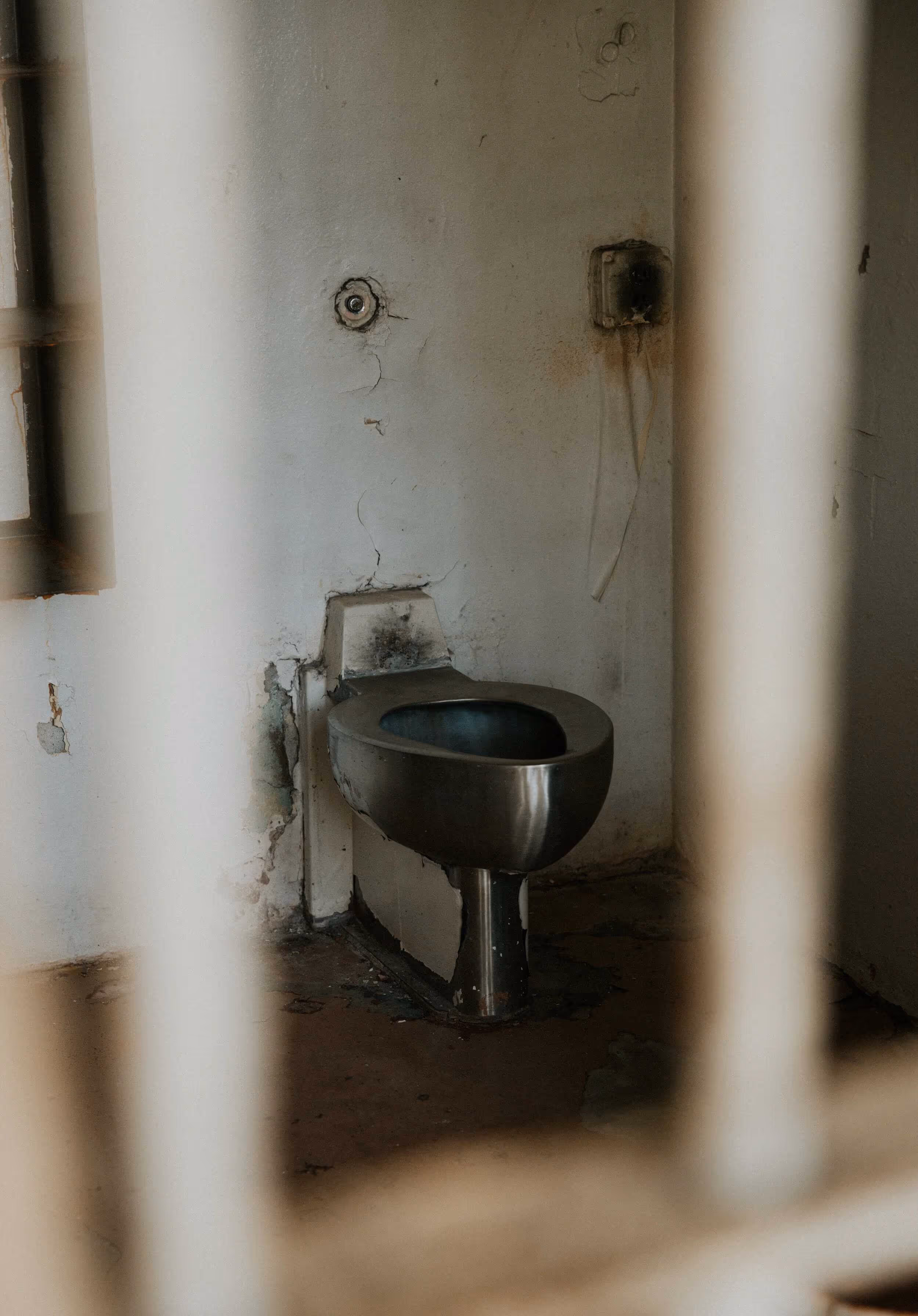 Rusty metal prison-style toilet attached to a stained, cracked wall, viewed through vertical prison bars.