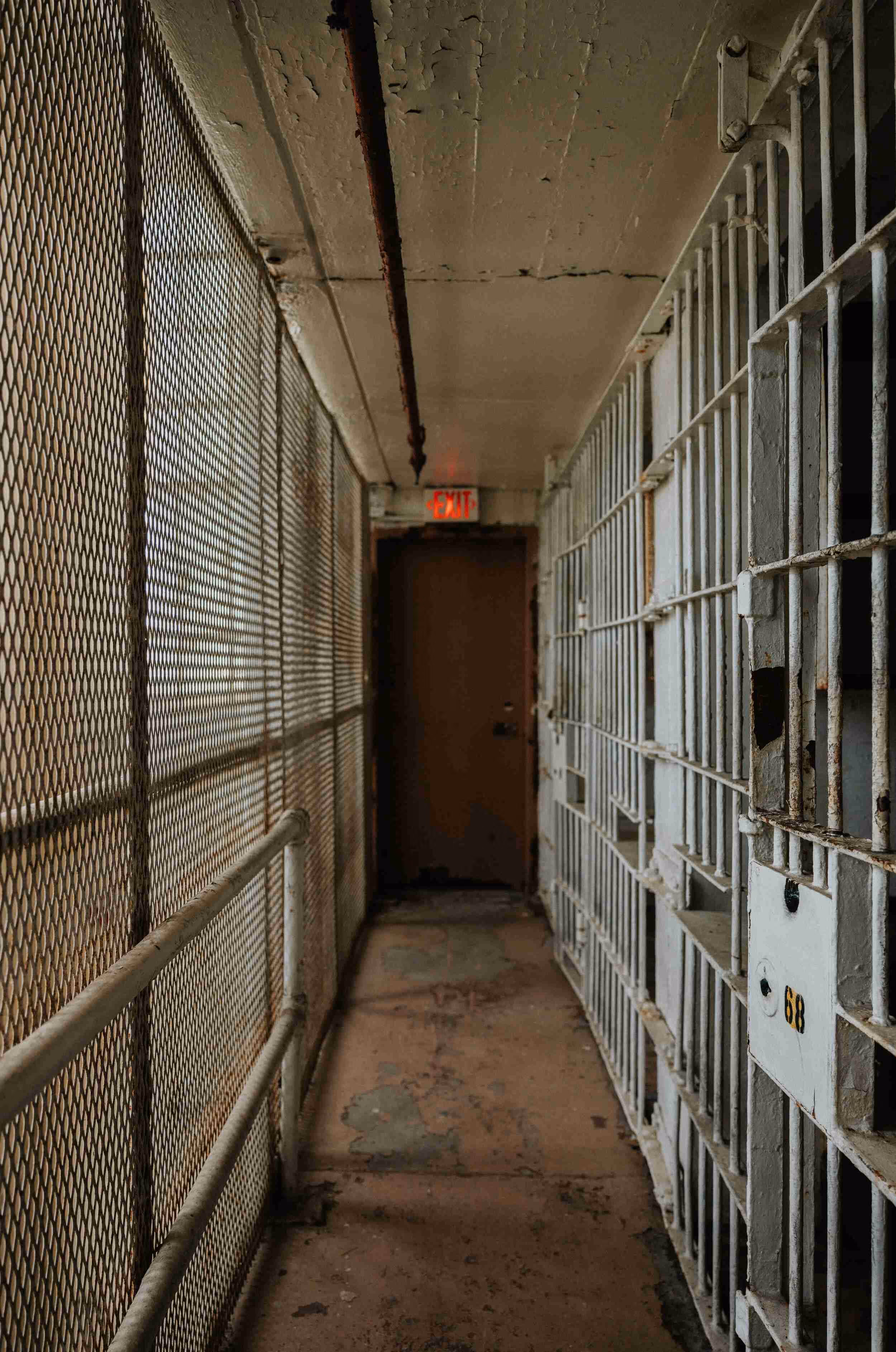 Narrow prison corridor with metal bars on the right, chain-link fence on the left, and an exit door at the end.