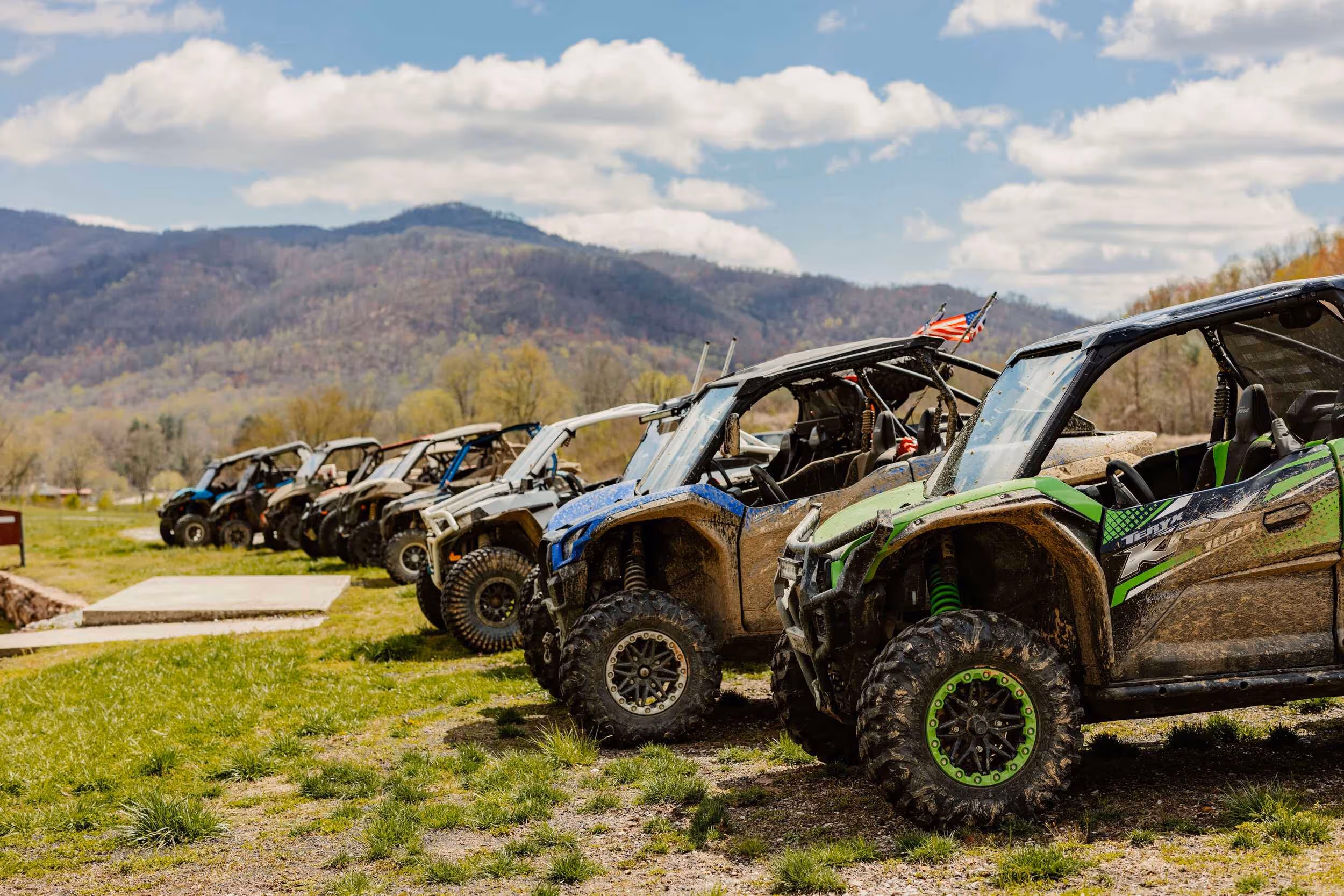 A row of muddy off-road side-by-side ATVs parked on grass with mountains and partly cloudy sky in the background.