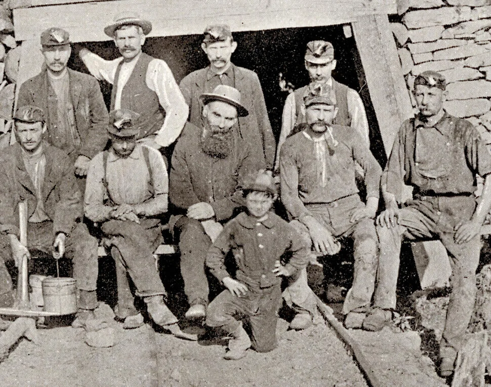 Black and white photo of ten coal miners and one boy posed outside a mine entrance, wearing work clothes and caps.