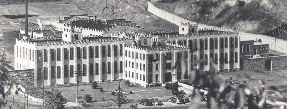Black and white historical aerial photo of the large, multi-winged Brushy penitentiary with battlement-style roof edges, surrounded by a fenced area and hills in the background.