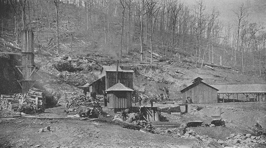 Black and white photo of a mining site with small wooden buildings, stacks of logs, and bare trees on a hillside in the background.