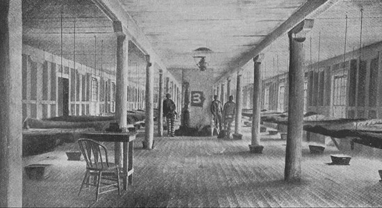 Black and white photo of a large, empty prison boarding room with rows of beds along both sides and two men standing in the middle near a stove.