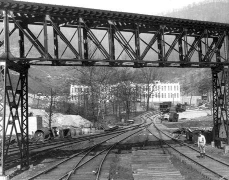 Black and white photo of a steel railway trestle bridge over multiple railroad tracks with a man walking near the tracks and buildings in the background.