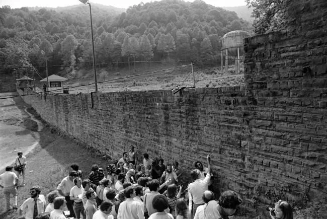 Black and white photo of a group of people gathered near the tall prison brick wall inside the Brushy Mountain Penitentiary where James Earl Ray and six other prisoners escaped.