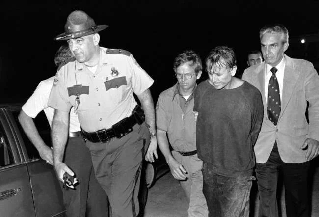 Police officers escorting a handcuffed James Earl Ray in casual clothes at night.