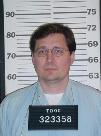 Mugshot of Byron Looper with glasses wearing a light blue shirt standing in front of a height measurement chart holding a TDOC placard with ID number 323358.