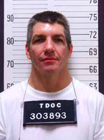 Mugshot of a Paul Dennis Reid with short dark hair wearing a white shirt and a TDOC identification placard with number 303893, standing against a height measurement chart.