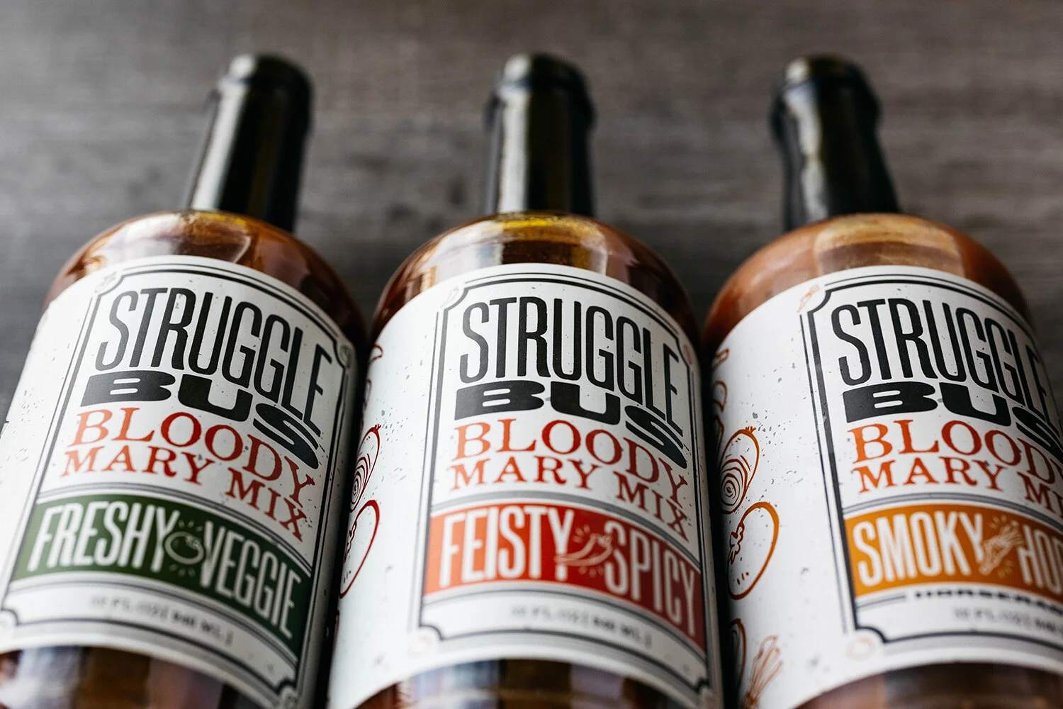 Three bottles of Struggle Bus Bloody Mary Mix in Fresh Veggie, Feisty Spicy, and Smoky Hot flavors on a dark wooden surface.