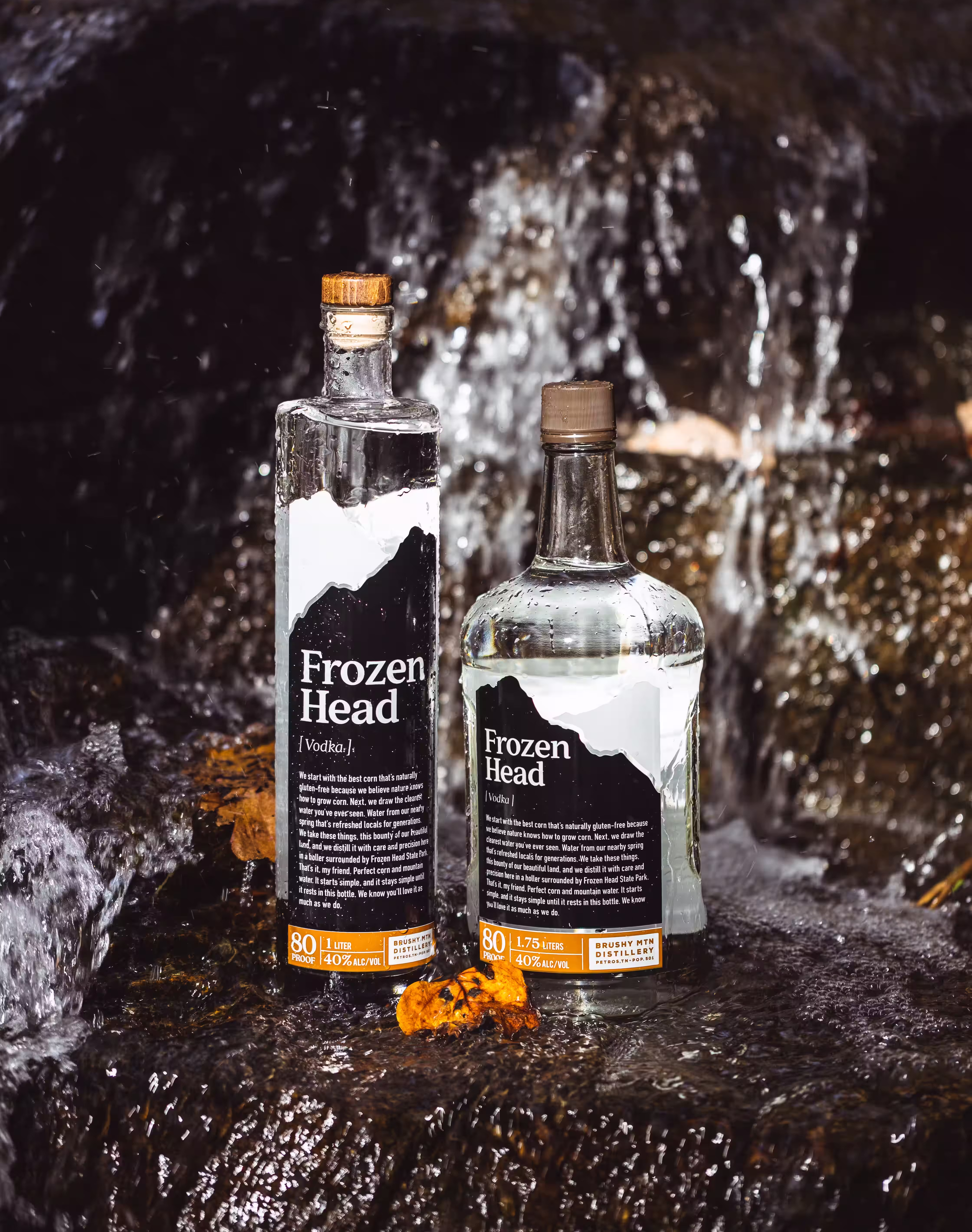 Two bottles of Frozen Head Vodka with water droplets on them, placed on a wet rock near a waterfall.