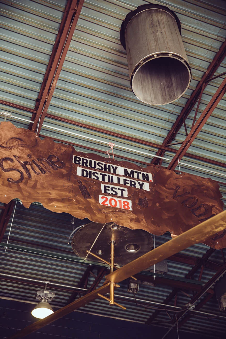 Rustic wooden sign hanging from a metal ceiling that reads 'Brushy Mtn. Distillery Est 2018' with the words 'Shine' and 'Lodi' carved on the sides.