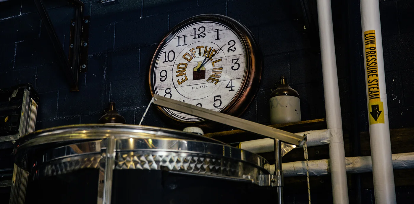 Industrial distillery room with a large clock reading 'End of the Line' and pipes labeled 'Low Pressure Steam'.
