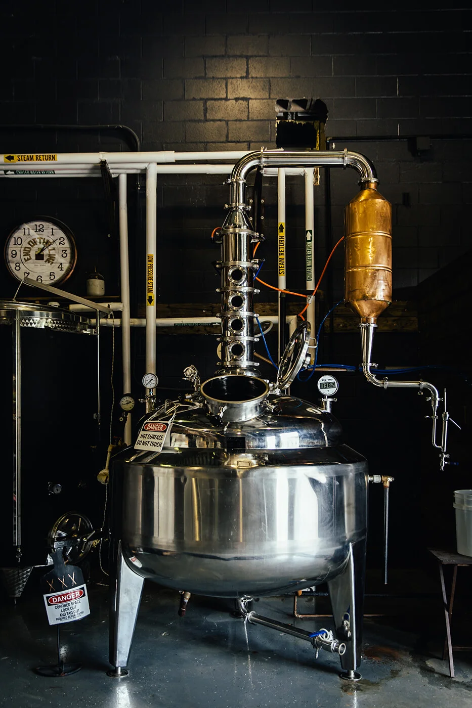 Stainless steel distillation equipment with pipes and gauges inside a distillery against a black brick wall.