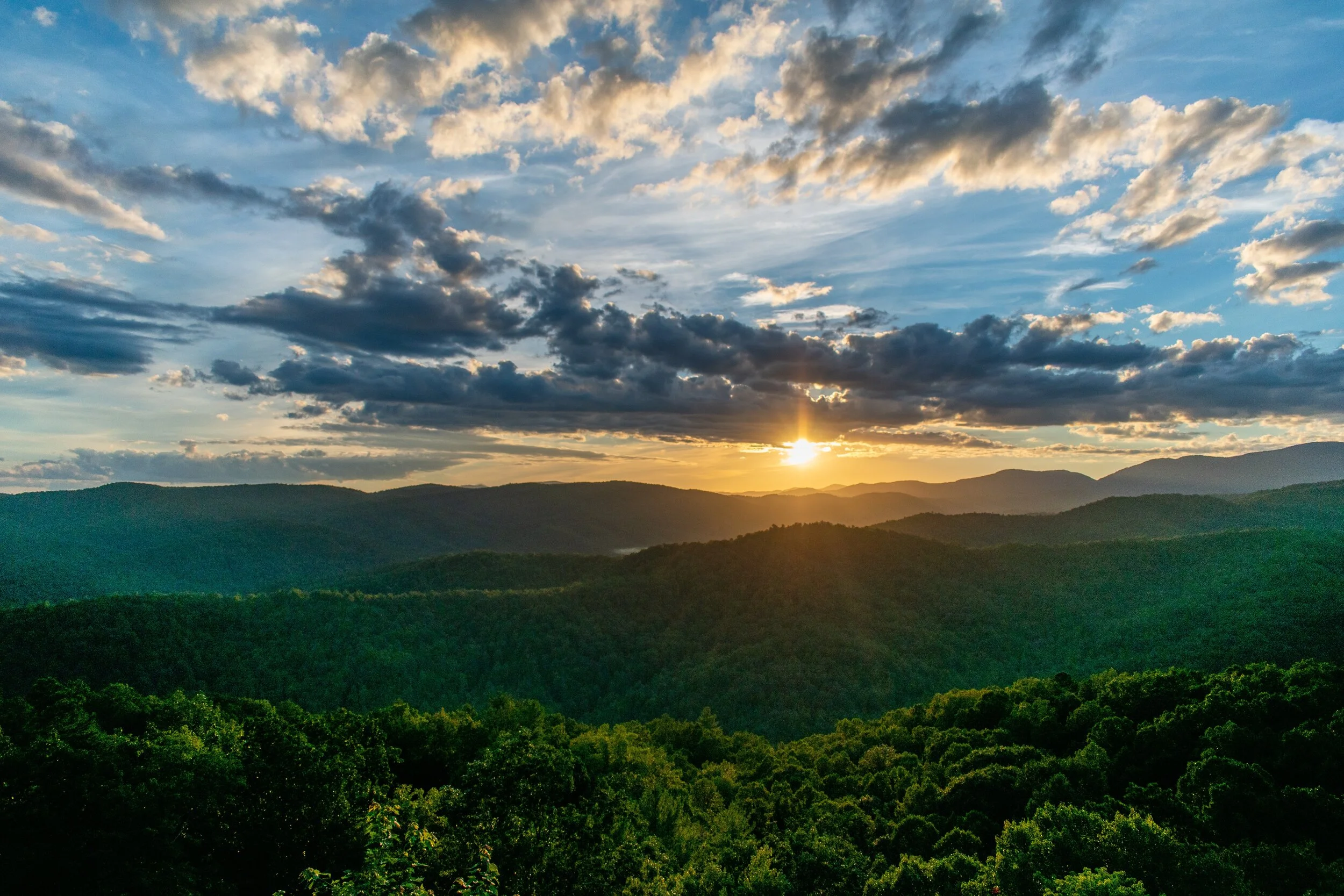 Sun setting over layered forested mountains under a partly cloudy blue sky.