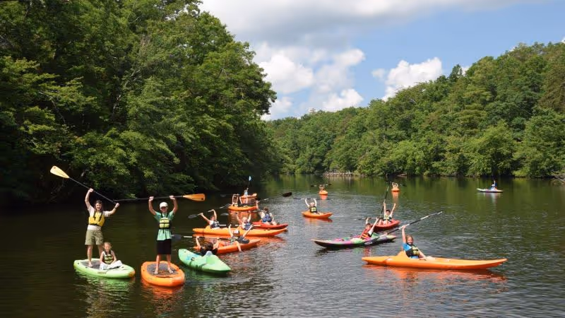 Group of people kayaking and paddleboarding on a calm river surrounded by green trees under a partly cloudy sky.