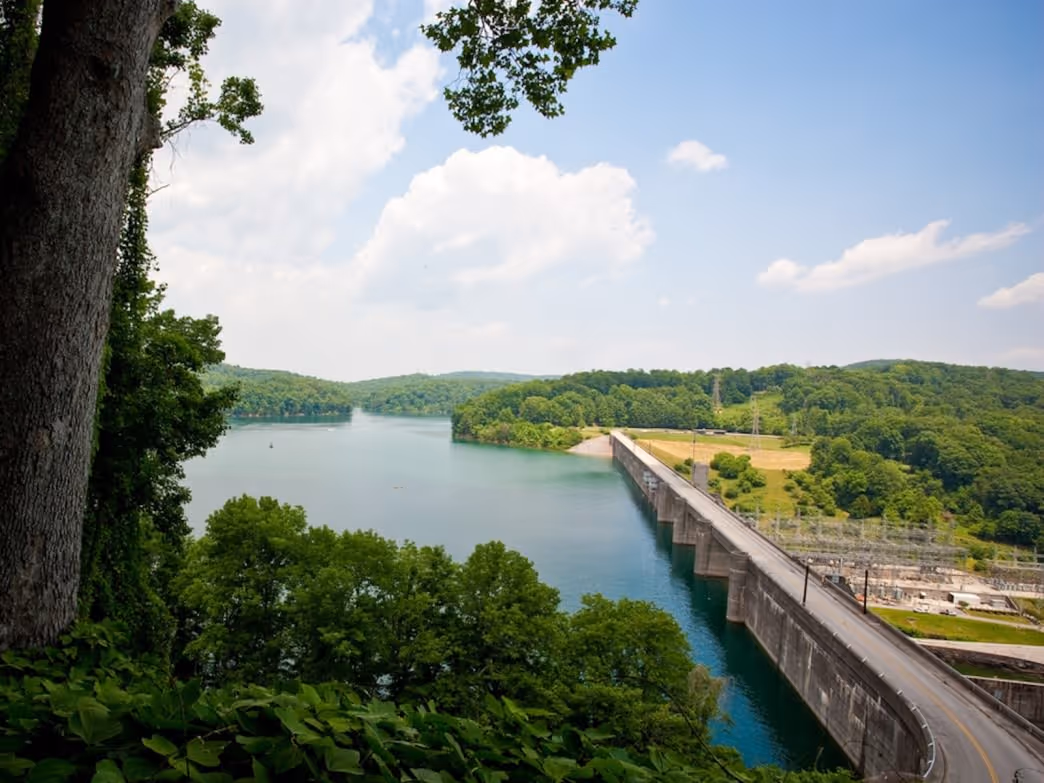 Wide view of a dam stretching across a river surrounded by green trees and hills under a partly cloudy sky.