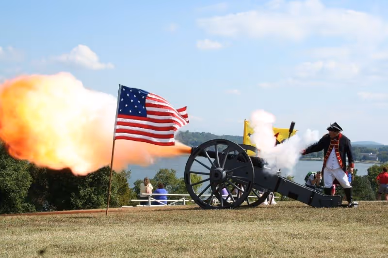 Historical reenactor in colonial uniform firing a cannon with a large flame near an American flag on a grassy hill overlooking a river.