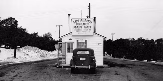 Black and white photo of a car at the entrance guard post labeled 'Los Alamos Project Main Gate' requiring passes for entry.