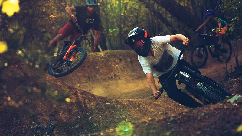 Three mountain bikers wearing helmets riding hard on a dirt trail through a forested area.