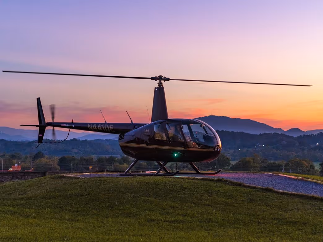 Black helicopter parked on grassy hill with mountains and sunset sky in the background.