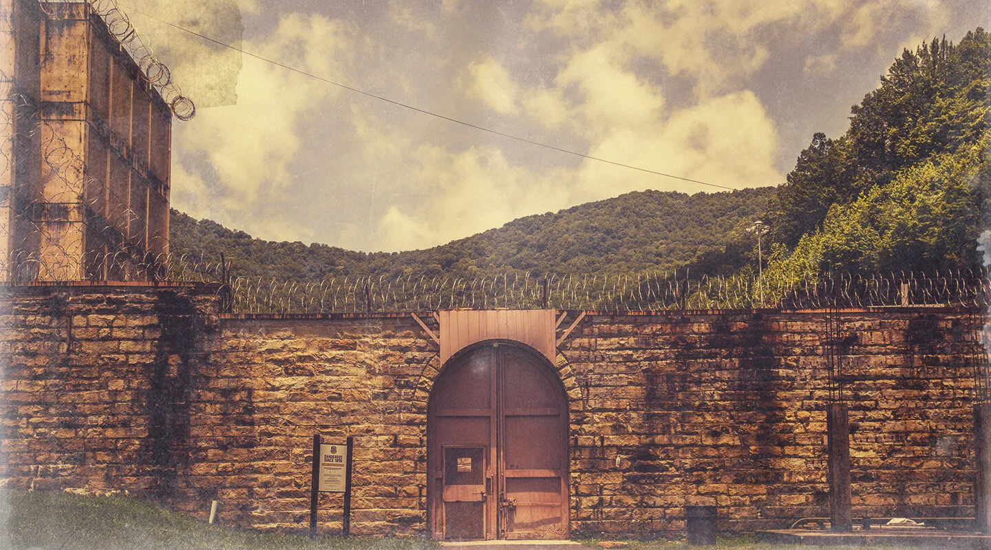 Stone prison wall topped with barbed wire and a large closed metal gate under a cloudy sky with forested hills in the background.