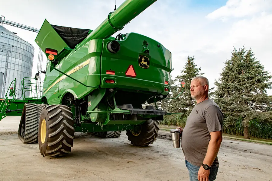 A farmer stands next to a harvester with grain bins in the background.