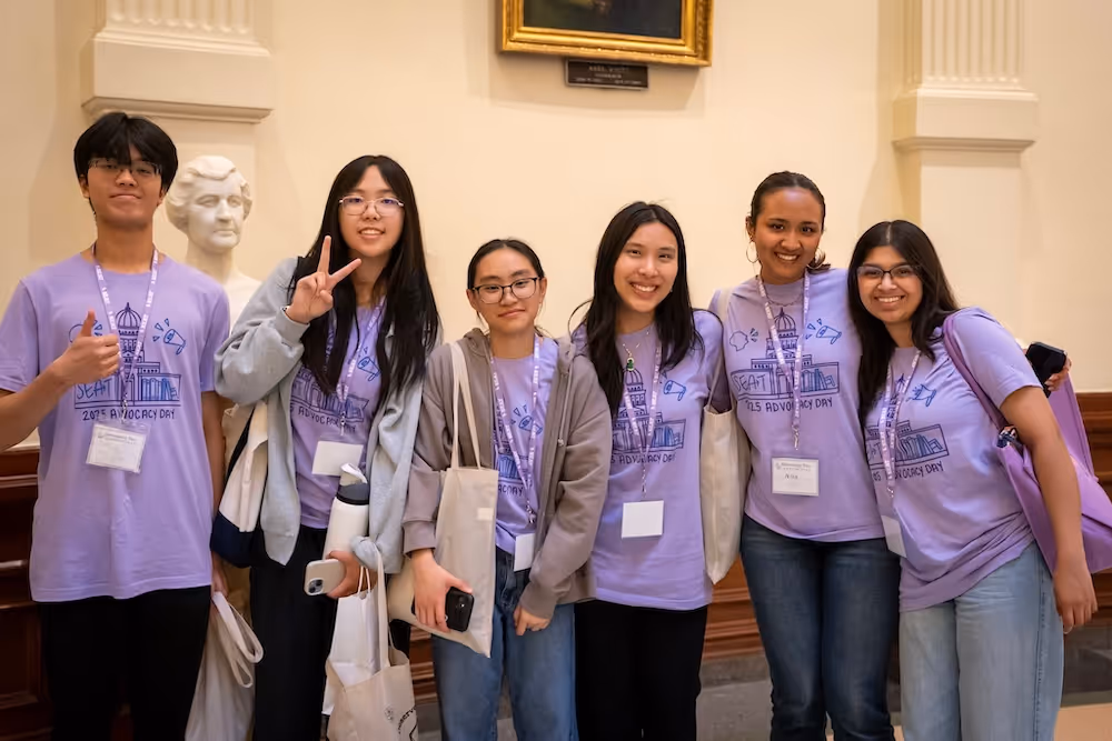 Group of six young people wearing matching purple t-shirts and name tags for SEAT Advocacy Day posing inside the Texas Capitol with smiles and casual gestures.