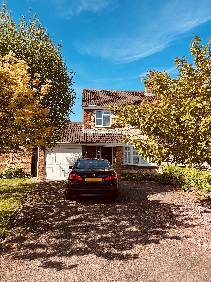 Black BMW car parked on a driveway in front of a two-story brick house with a white garage door, surrounded by green and yellow trees under a blue sky.