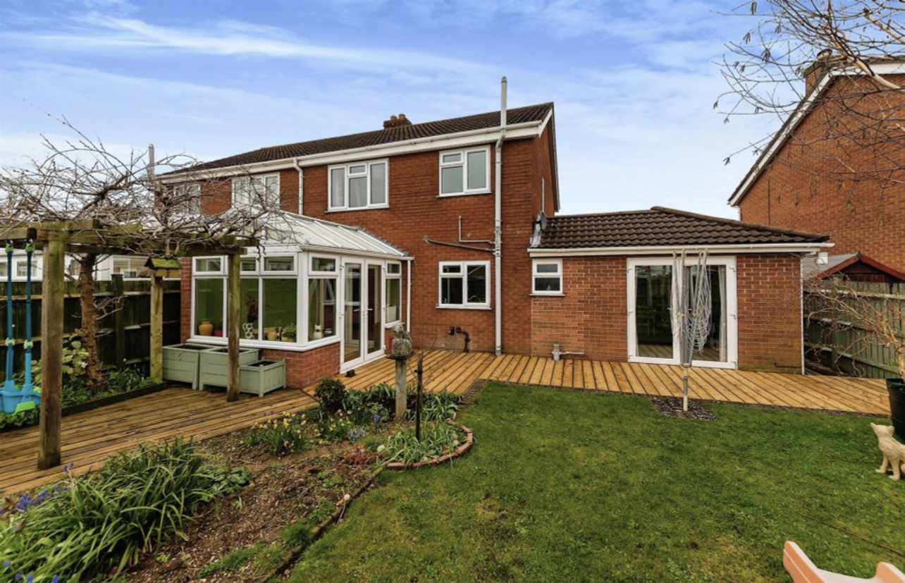 Backyard of a brick two-story house with a wooden deck, conservatory, garden bed, and a lawn under a blue sky.