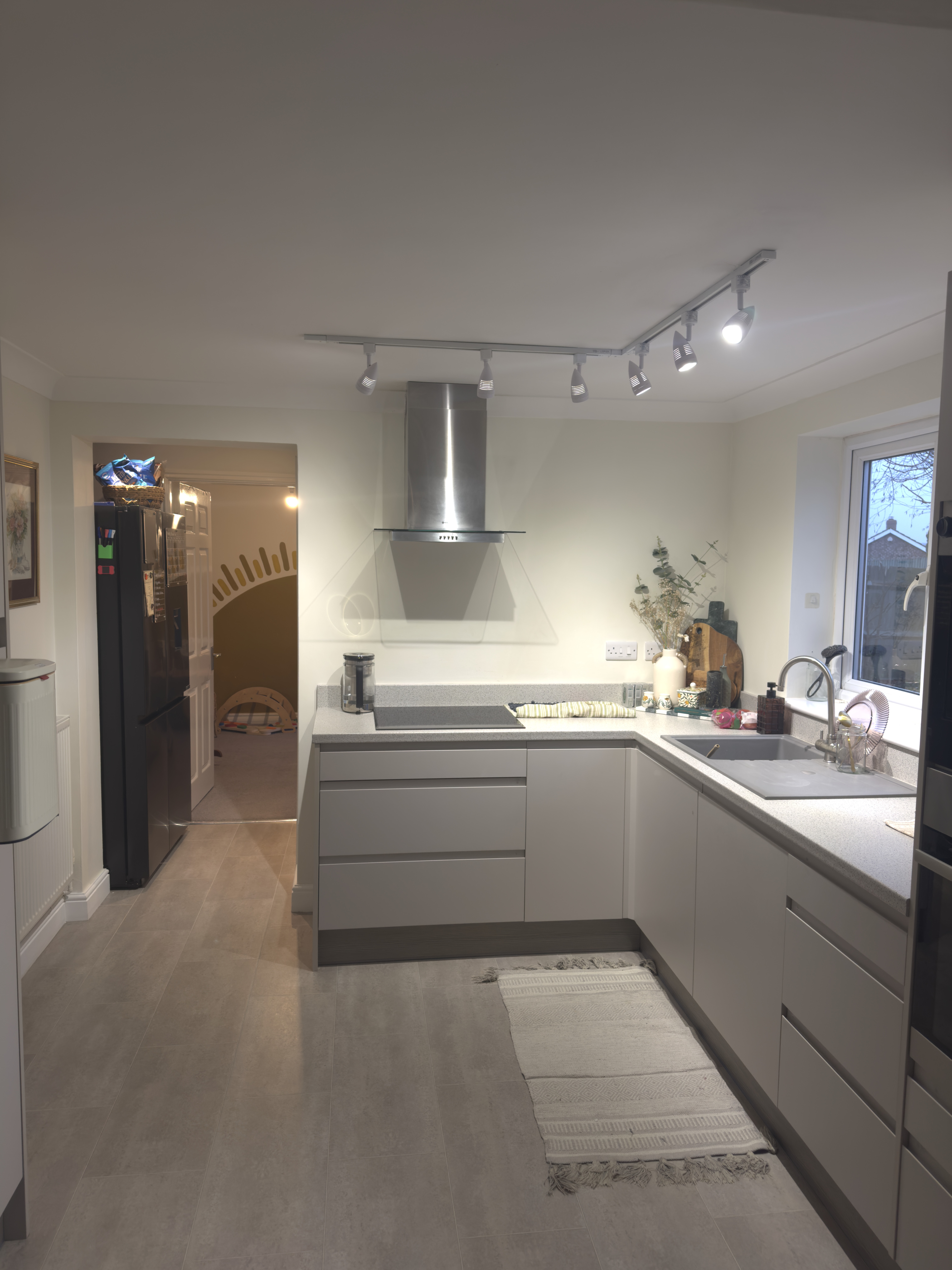 Modern kitchen with light gray cabinets, steel range hood, and a window above the sink, illuminated by overhead track lighting.