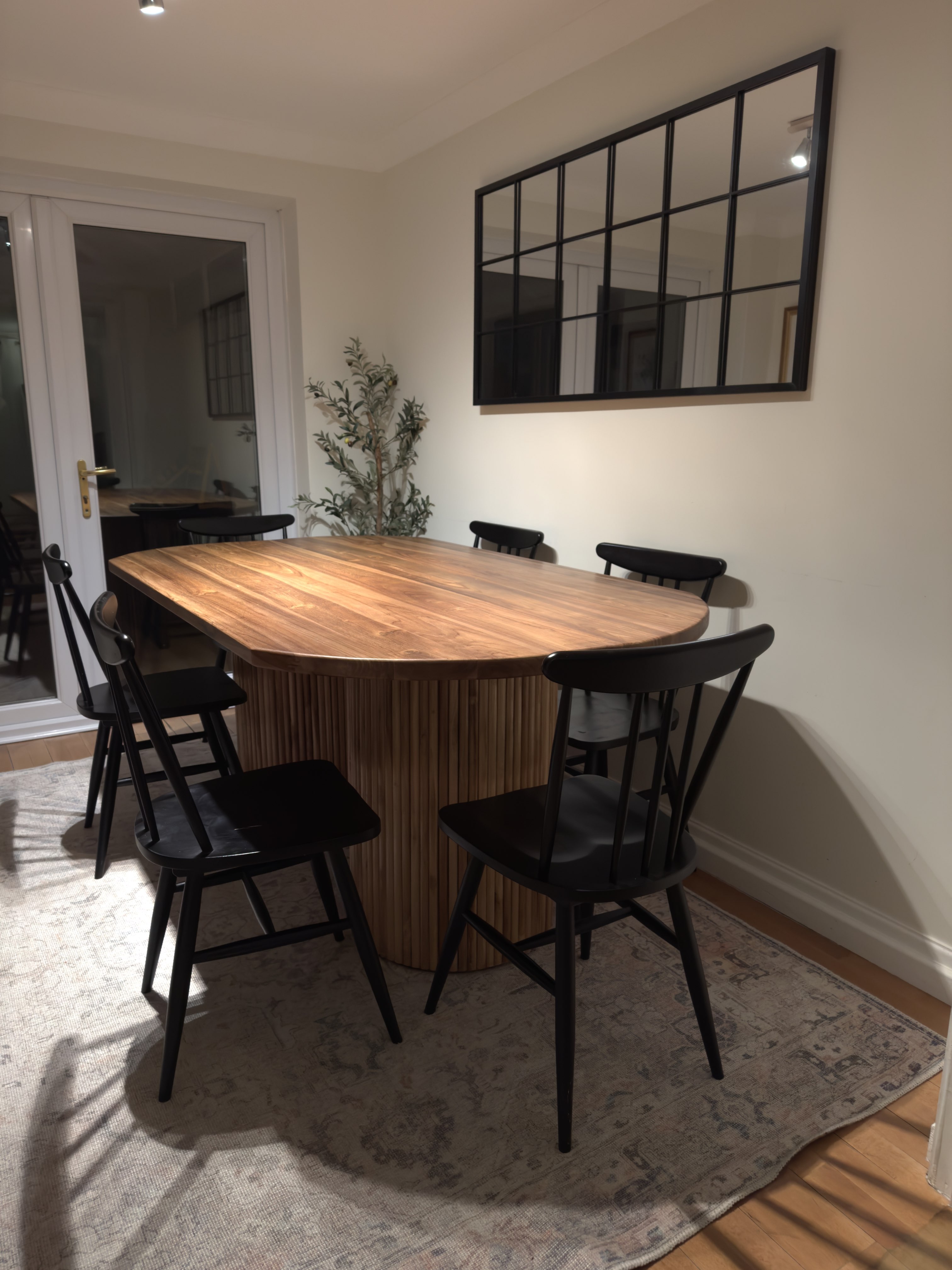 Modern dining area with a wooden oval table surrounded by six black chairs, a patterned rug underneath, a plant in the corner, and a large black-framed grid mirror on the wall.