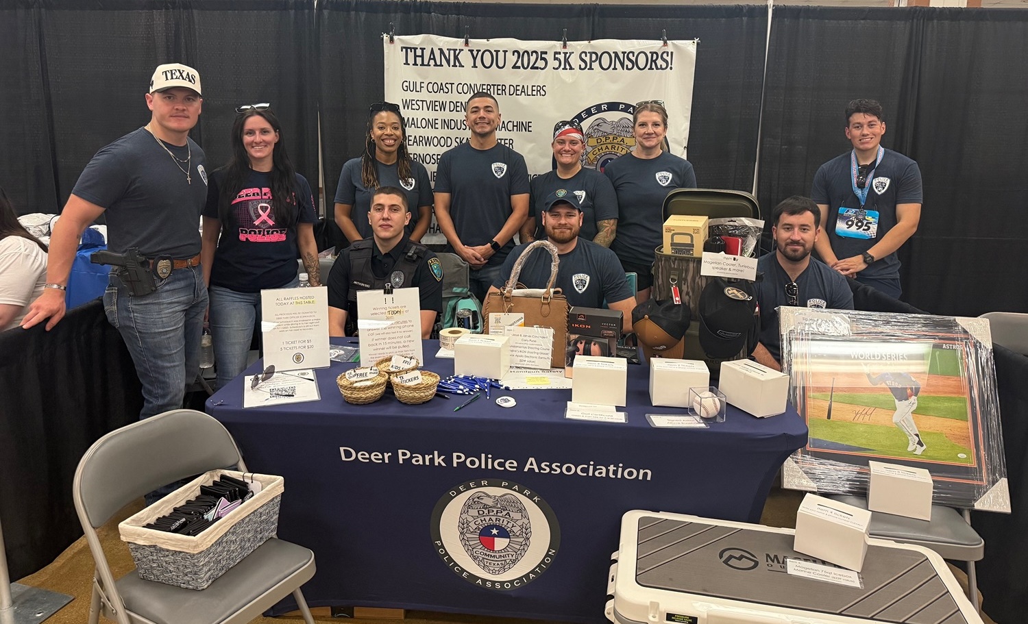 Group of Deer Park Police Association members standing and sitting behind a booth with raffle prizes and a thank you banner for 2025 5K sponsors.