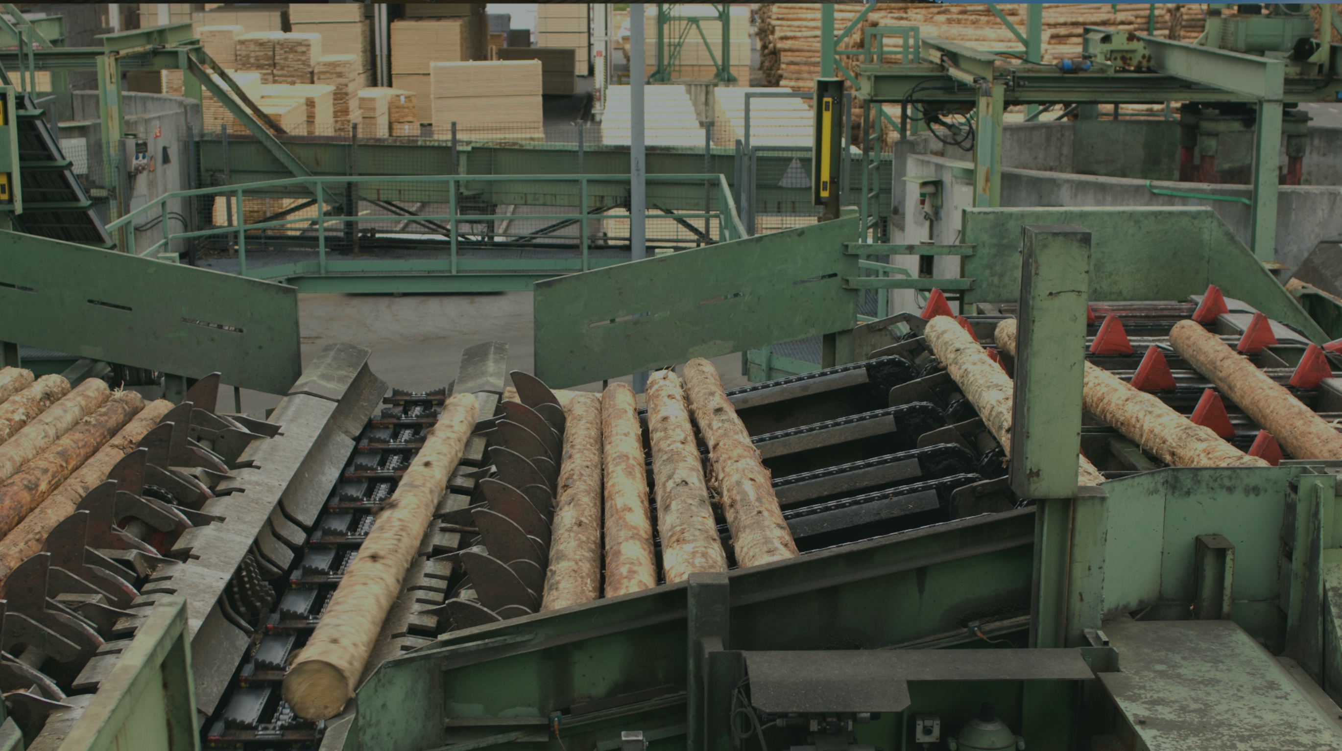 Industrial sawmill machinery processing logs with stacked wood planks in the background.