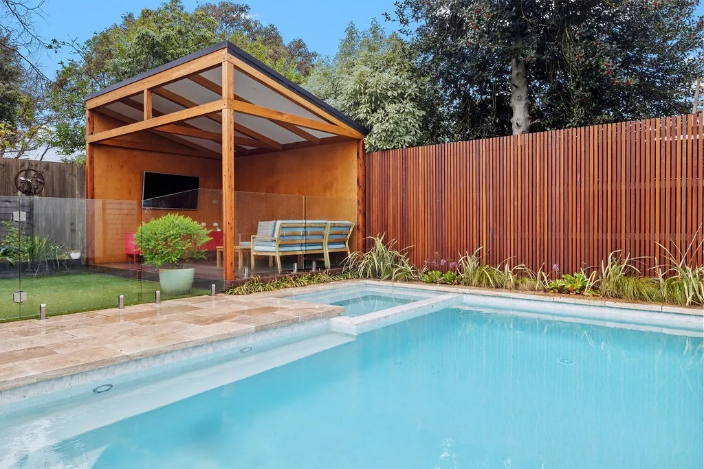 Backyard with a swimming pool, glass safety fence, wooden pergola with seating area, and tall wooden fence with greenery.